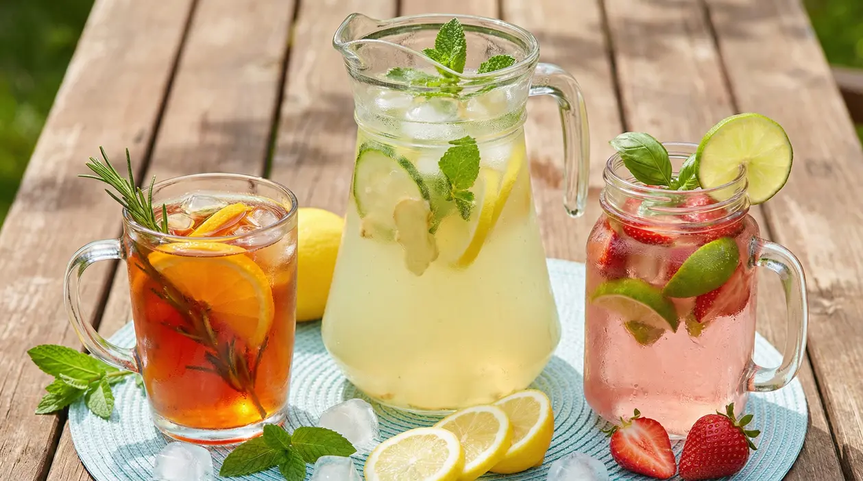 Tre bevande fresche in bicchieri di vetro con agrumi, menta e frutta su un tavolo di legno all’aperto