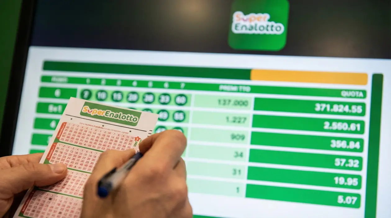 Una persona compila una schedina del SuperEnalotto davanti a uno schermo con i risultati.